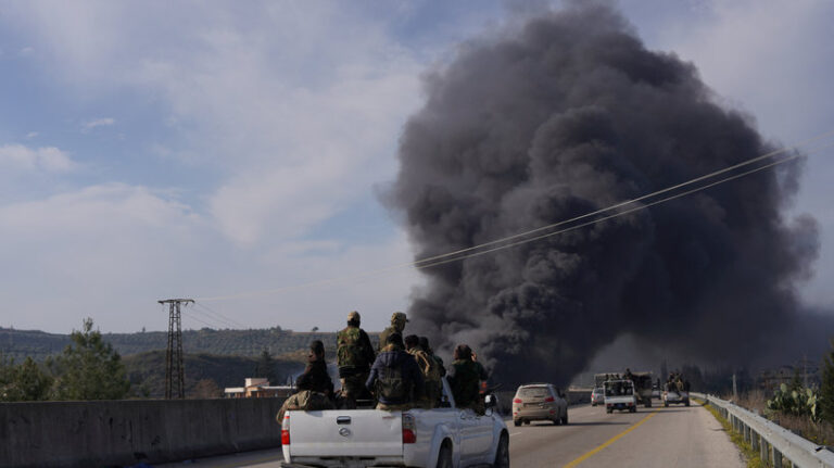 Smoke rises while members of the Syrian forces ride on a vehicle in Latakia